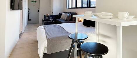 Una habitación de hotel moderna con una cama, una pequeña mesa de comedor, dos taburetes y un ventanal amplio.