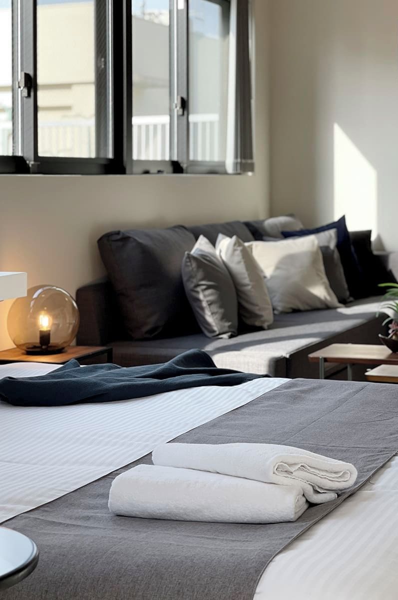 Une chambre d’hôtel moderne, équipée d’un lit, d’une table de chevet avec une lampe, d’une fenêtre avec des stores et offrant une vue sur l’extérieur.