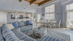 Ampla sala de estar com um sofá grande, uma mesa de jantar e cozinha ao fundo.