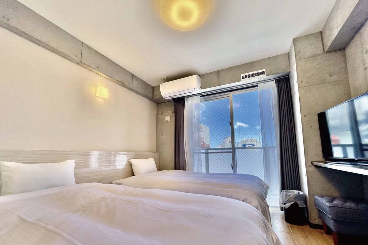 Habitación de hotel con dos camas, un ventanal con vistas, un equipo de aire acondicionado y un televisor a la derecha.