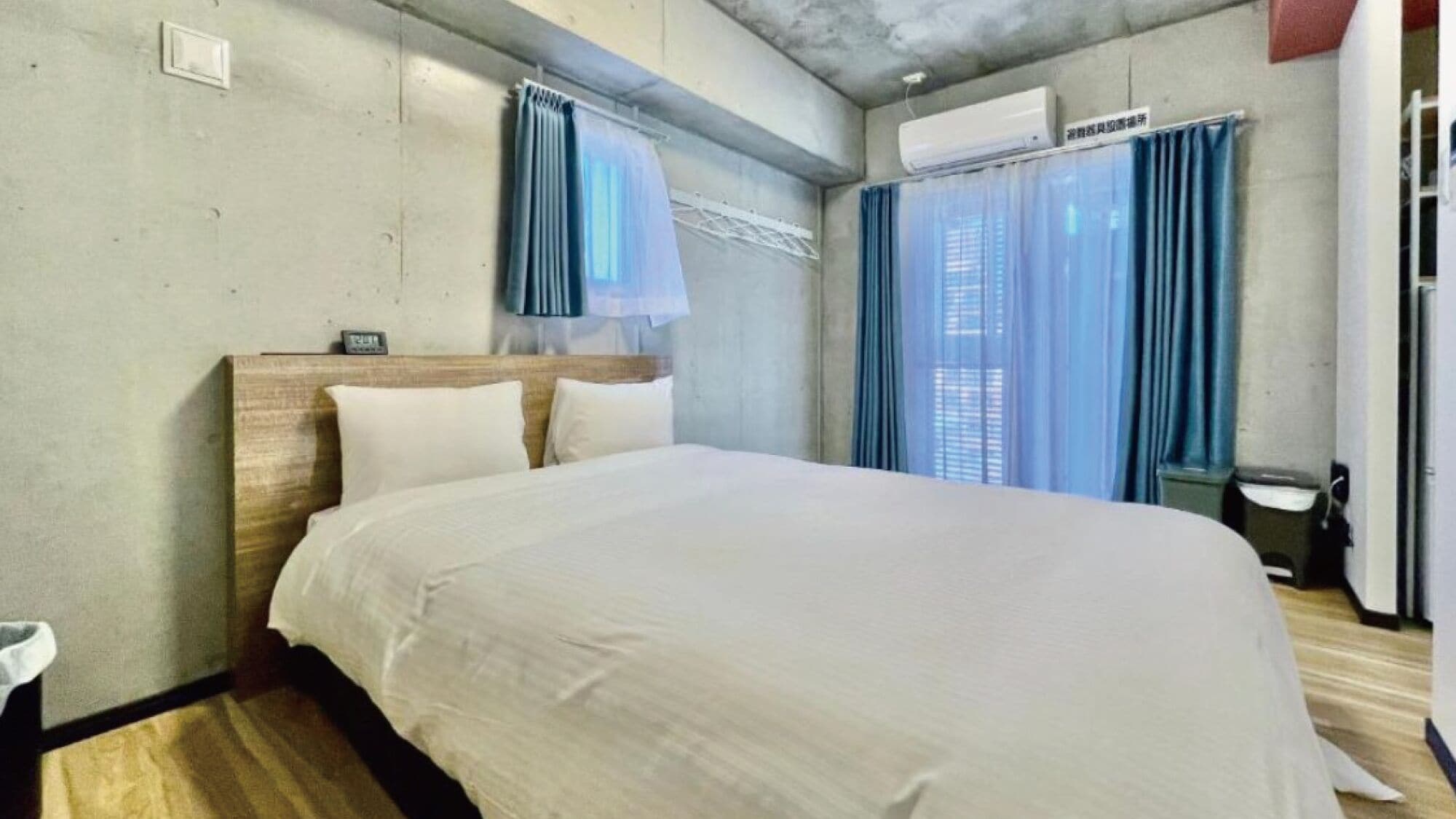 Een hotelkamer met een bed, een houten hoofdeinde, een wandairconditioner en een raam met blauwe gordijnen.