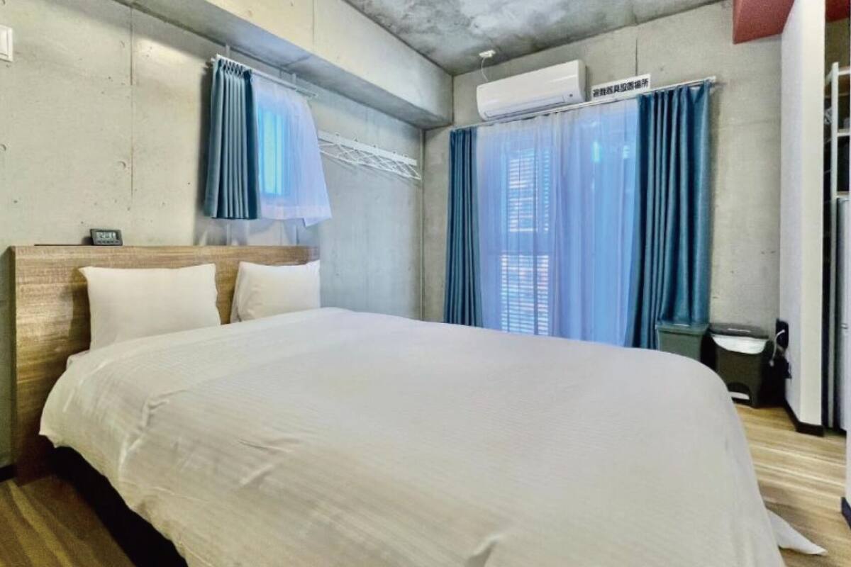 Una habitación de hotel con una cama, cabecera de madera, aire acondicionado de pared y una ventana con cortinas azules.