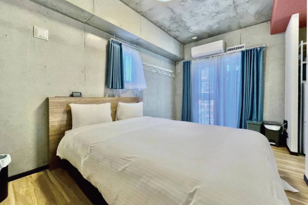 Habitación de hotel con cama, cortinas, unidad de aire acondicionado y suelo de madera.