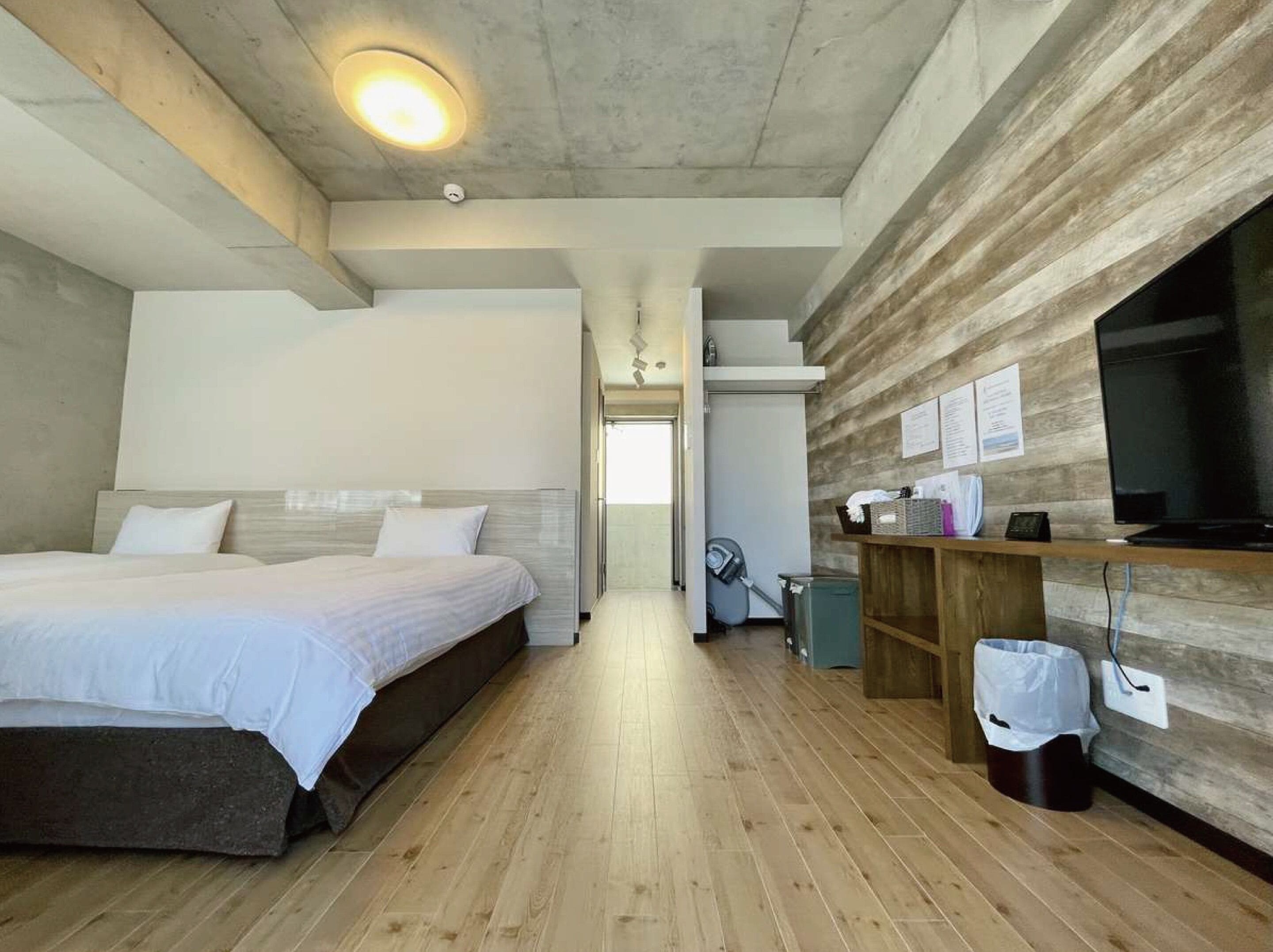 Une chambre d’hôtel moderne avec un grand lit, un plancher en bois, un bureau avec une télévision et une petite poubelle.