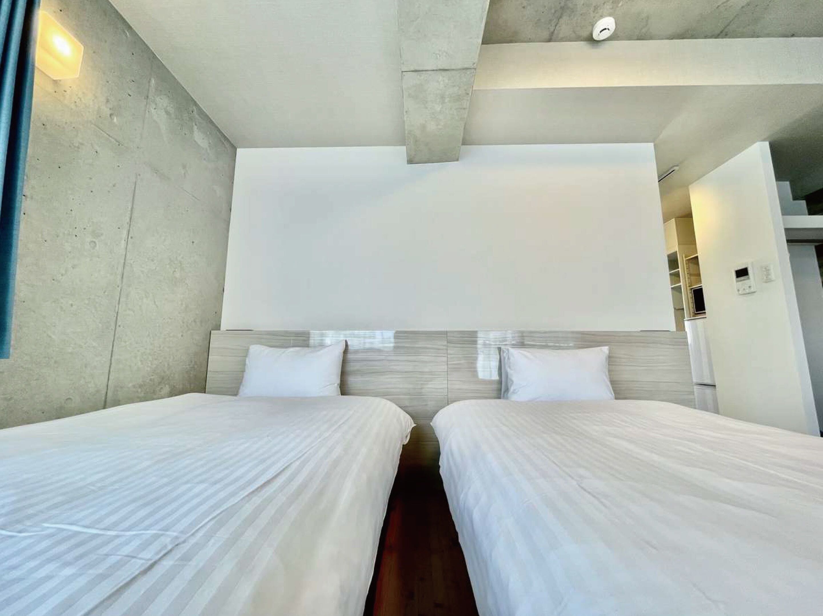 A modern hotel room with two beds, a concrete accent wall, and a large window.
