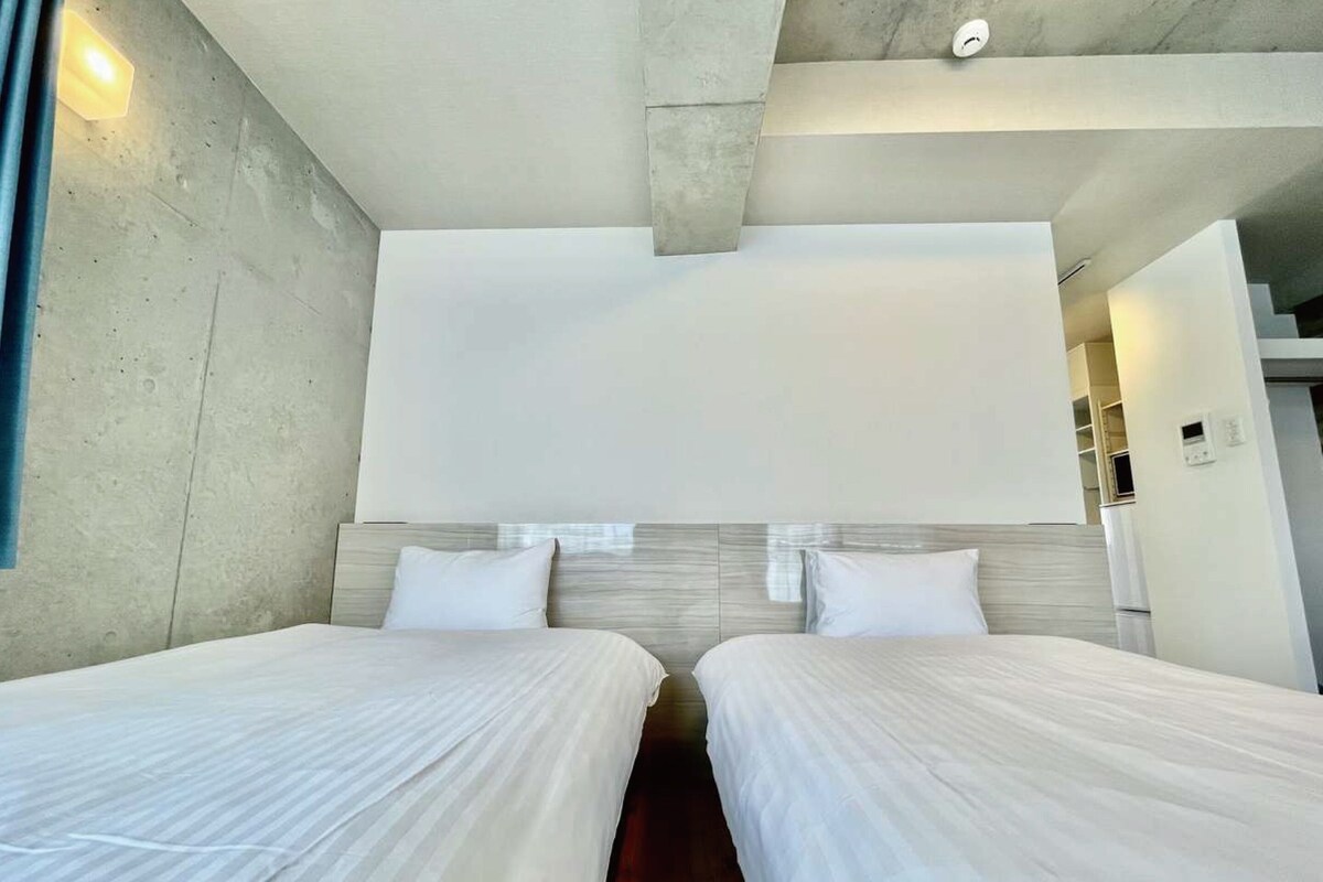 A modern hotel room with two beds, a concrete accent wall, and a large window.
