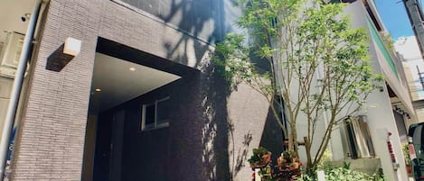 A modern building with a brick facade, a covered entrance, and a small garden area with plants and a tree.