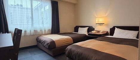 A hotel room with two beds, a desk, a chair, a lamp, and a window with curtains.