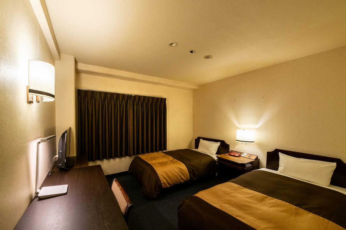 Ein Hotelzimmer mit zwei Betten, einem Schreibtisch mit Computer, einer Lampe und einem Fenster mit Vorhängen.