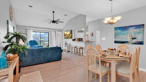 Un salon moderne avec un canapé bleu, une table à manger en bois et des chaises. On y trouve un ventilateur de plafond, un téléviseur et un tableau accroché au mur.