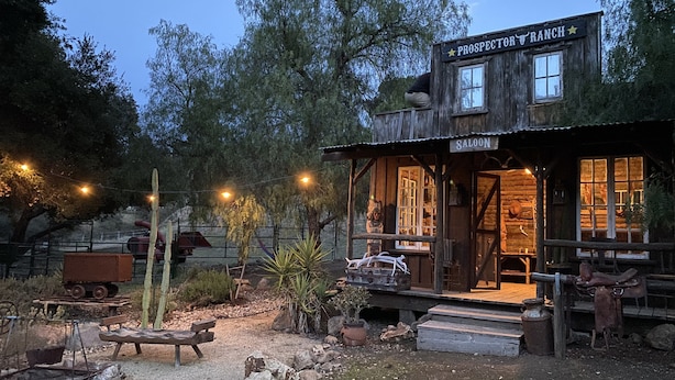 Een rustiek houten gebouw met de naam 'Prospector Ranch', voorzien van een veranda en een bord waarop 'Saloon' staat.