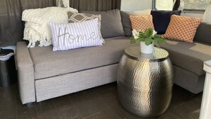 Een moderne woonkamer met een grijze bank, decoratieve kussens, een metalen bijzettafeltje en een raam met gordijnen.