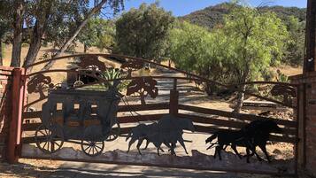 Una reja rústica con un carruaje de madera y siluetas de caballos, que da paso a un paisaje seco y montañoso.