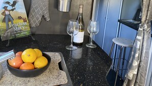 Een keuken met een zwart aanrecht, een hangende kruidenrek, een fles wijn, twee wijnglazen, een schaal met fruit en een kookboek.