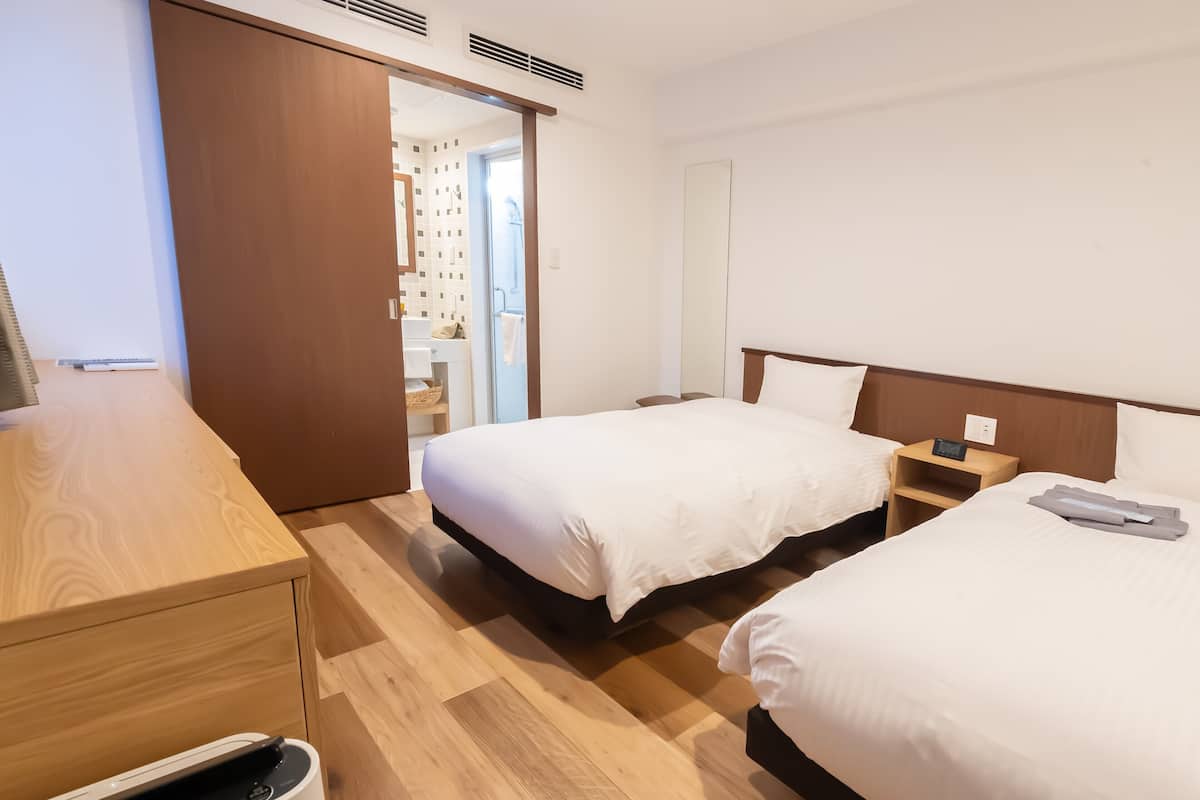 A hotel room with two beds, a wooden desk, a wardrobe, and a bathroom visible through an open door.