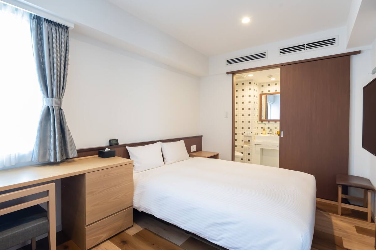 A modern hotel room with a wooden desk, a bed with white bedding, a small bedside table, a nightstand, a built-in wardrobe, and a bathroom with a tiled wall and a mirror.