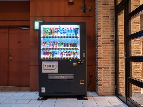 Máquina expendedora con diversas bebidas y aperitivos, ubicada frente a un muro de ladrillo y una puerta con la leyenda “非常口”.