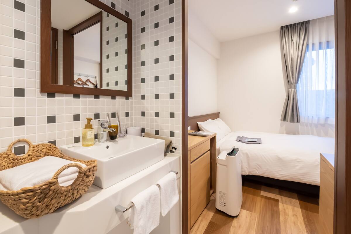A hotel room with a large mirror, a white sink, a wooden bedside table, a bed with white bedding, and a window with curtains.