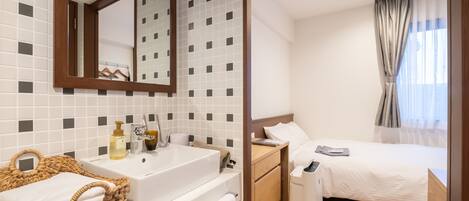 A hotel room with a large mirror, a white sink, a wooden bedside table, a bed with white bedding, and a window with curtains.