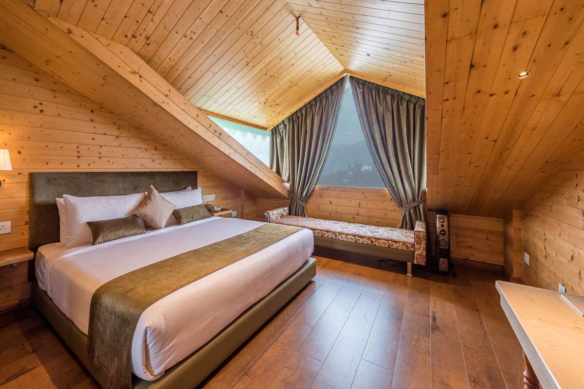 A cozy attic bedroom with a large bed, a bench, and a view through a window.