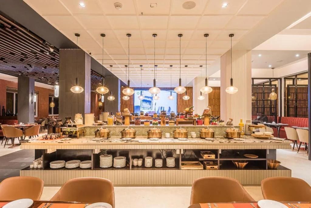A modern restaurant with a central bar area, multiple hanging pendant lights, and a variety of dishes displayed on the counter.