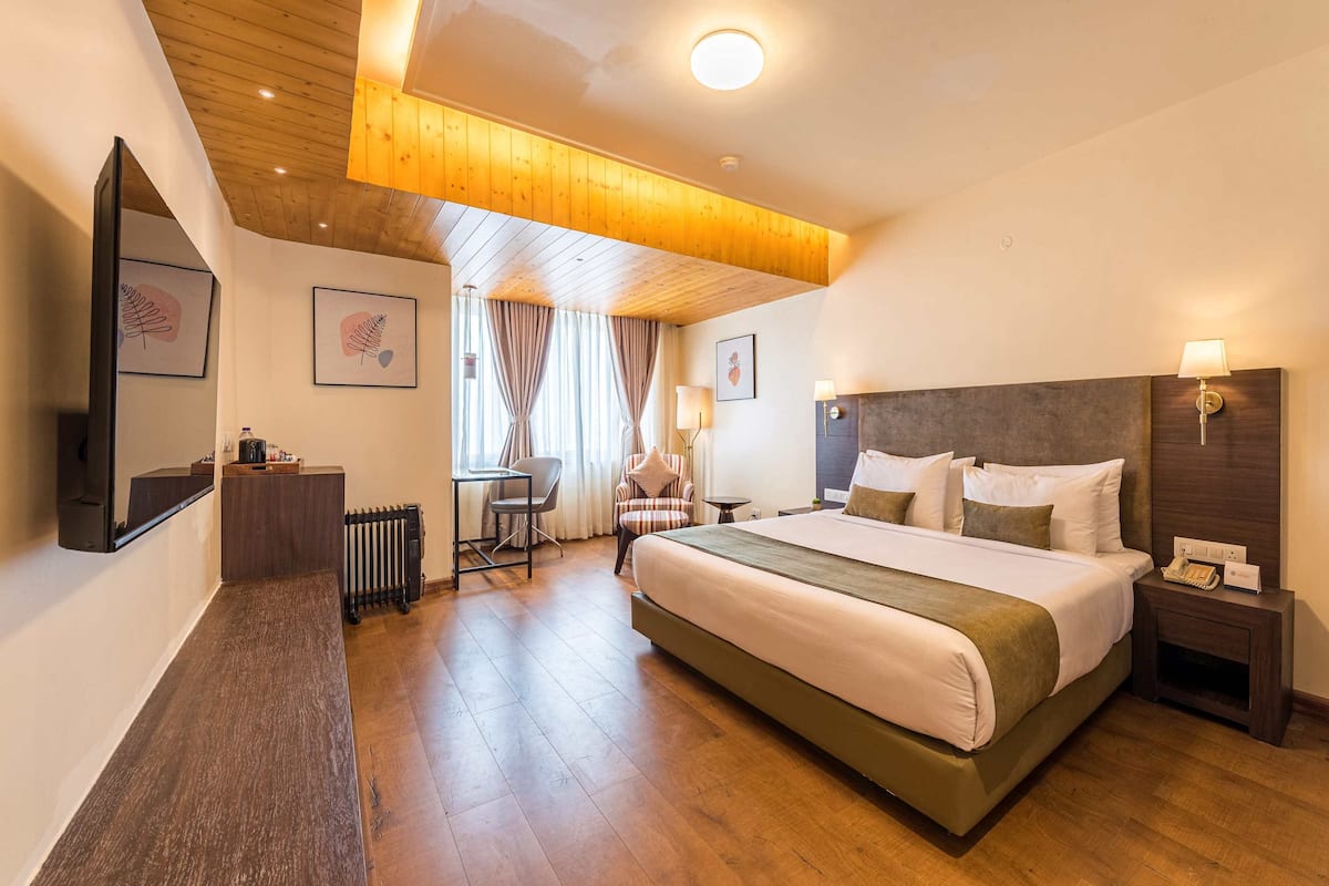 A hotel room with a large bed, a desk, a chair, a small table, a nightstand, and a mirror.