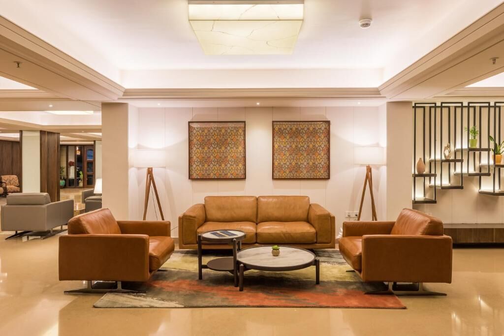 A spacious lobby with a brown leather sofa set, a coffee table, and two armchairs. There are two framed artworks on the wall and a potted plant on a shelf.