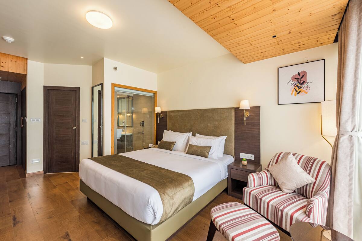 A hotel room with a large bed, a wooden ceiling, a striped armchair, and a framed picture on the wall.
