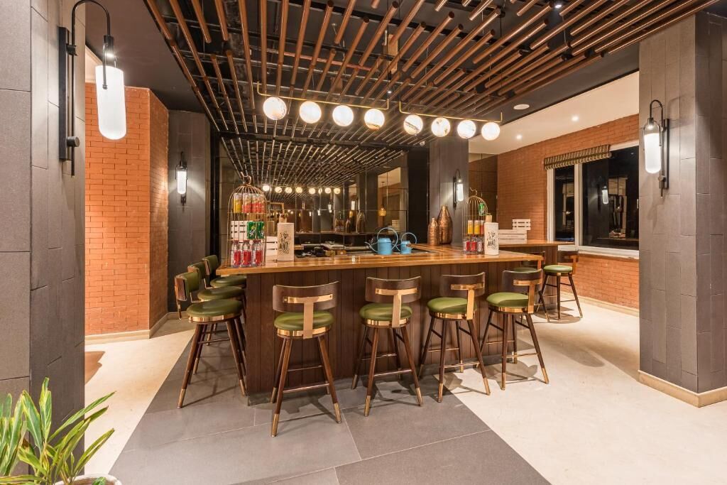 A modern bar area with a wooden bar counter, green cushioned stools, and a brick accent wall.