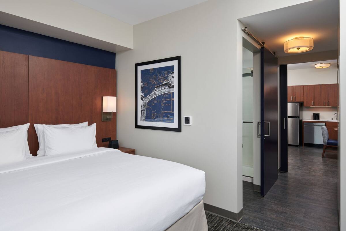 A hotel room with a large bed, a nightstand, a framed map on the wall, and a view into the kitchen area.