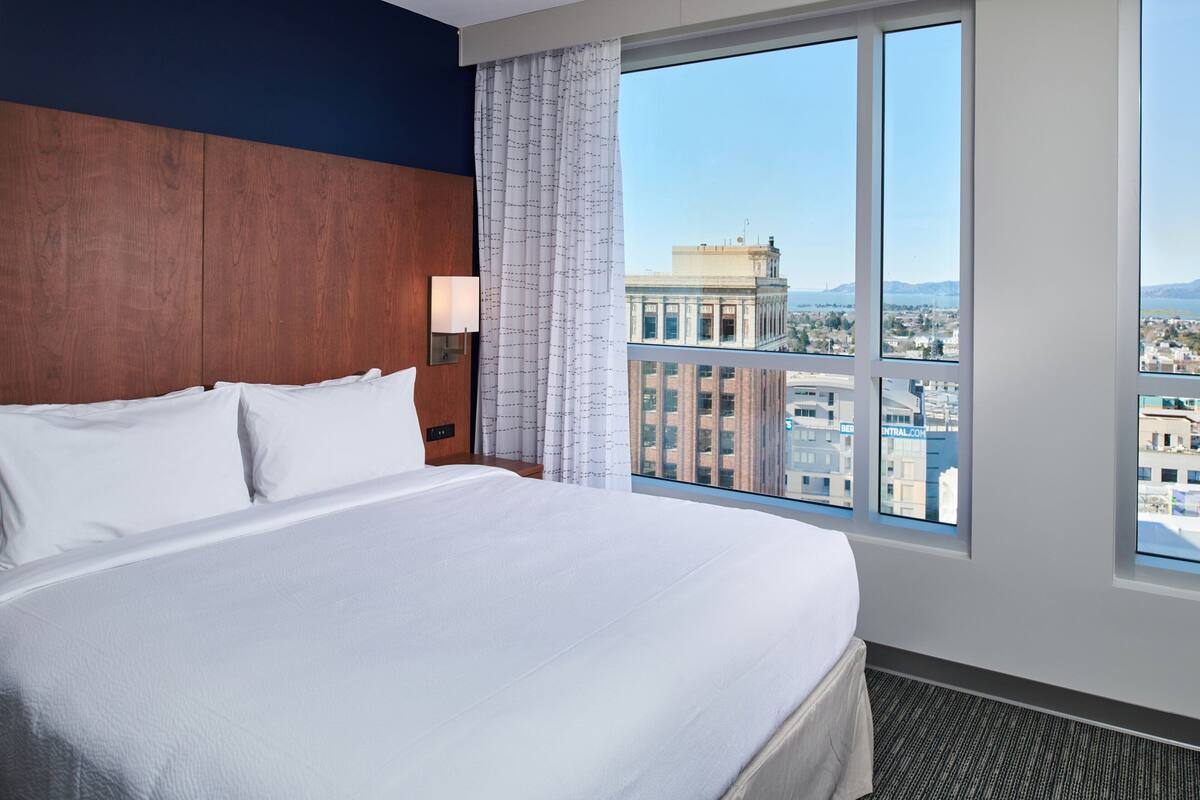 A hotel room with a large bed, a nightstand, a lamp, and a window with a city view.