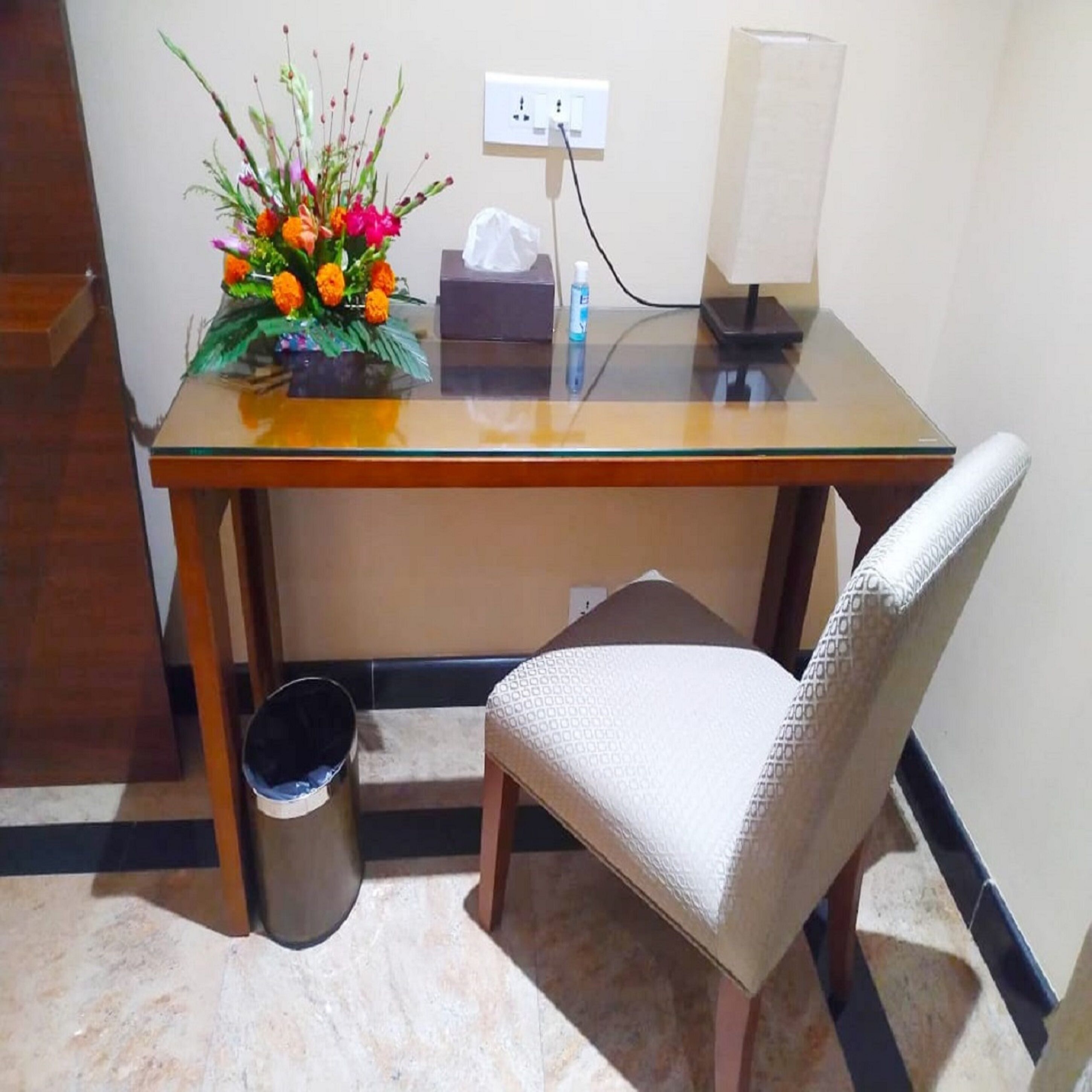 A small office space with a glass desk, a chair, a trash bin, a lamp, and a floral arrangement.