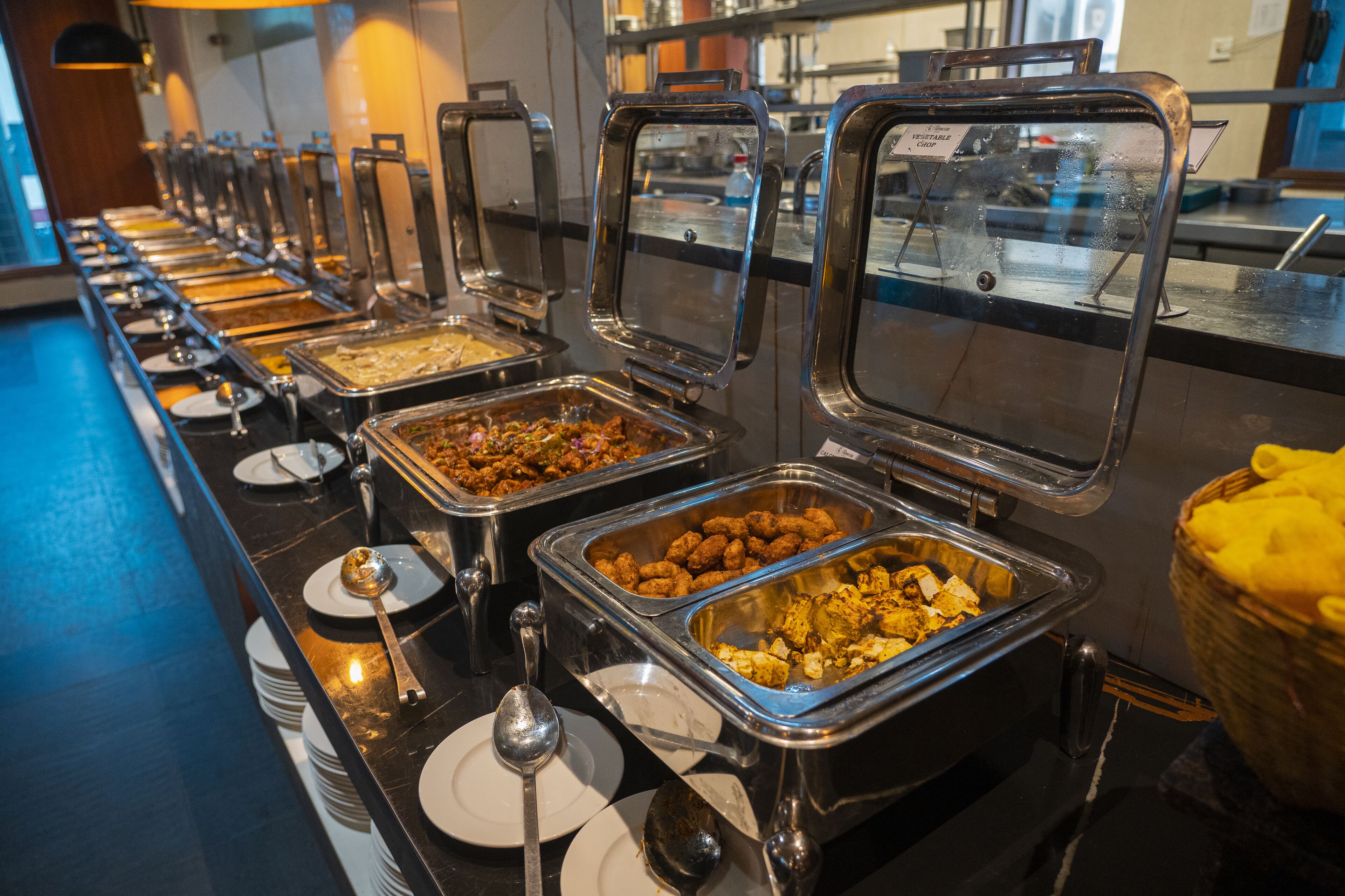 Un buffet proposant une variété de plats présentés dans des récipients en métal, disposés sur un comptoir avec des assiettes et des cuillères.