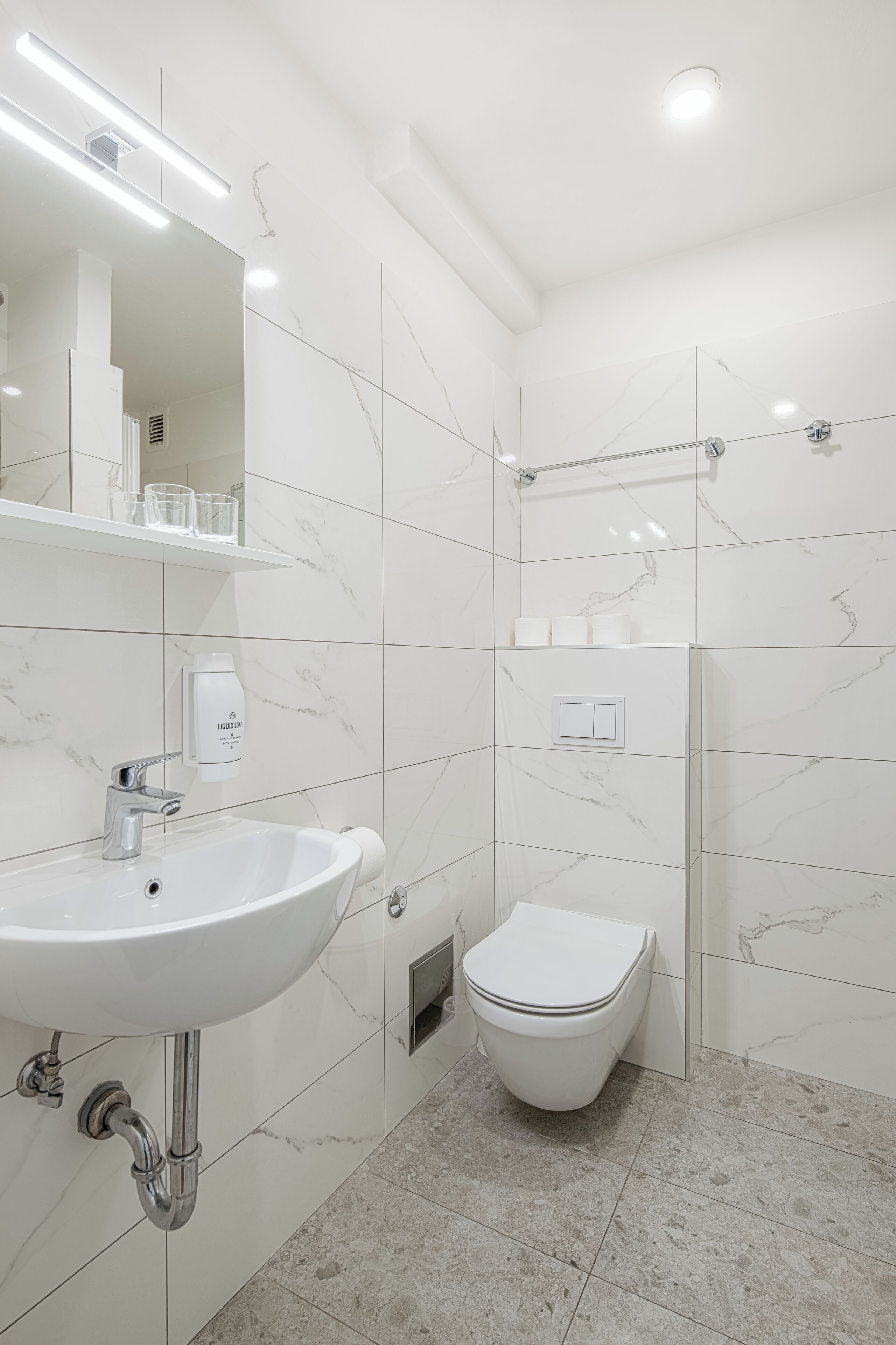 Un bagno moderno con lavandino, specchio e WC bianchi, piastrelle in marmo e zona doccia.