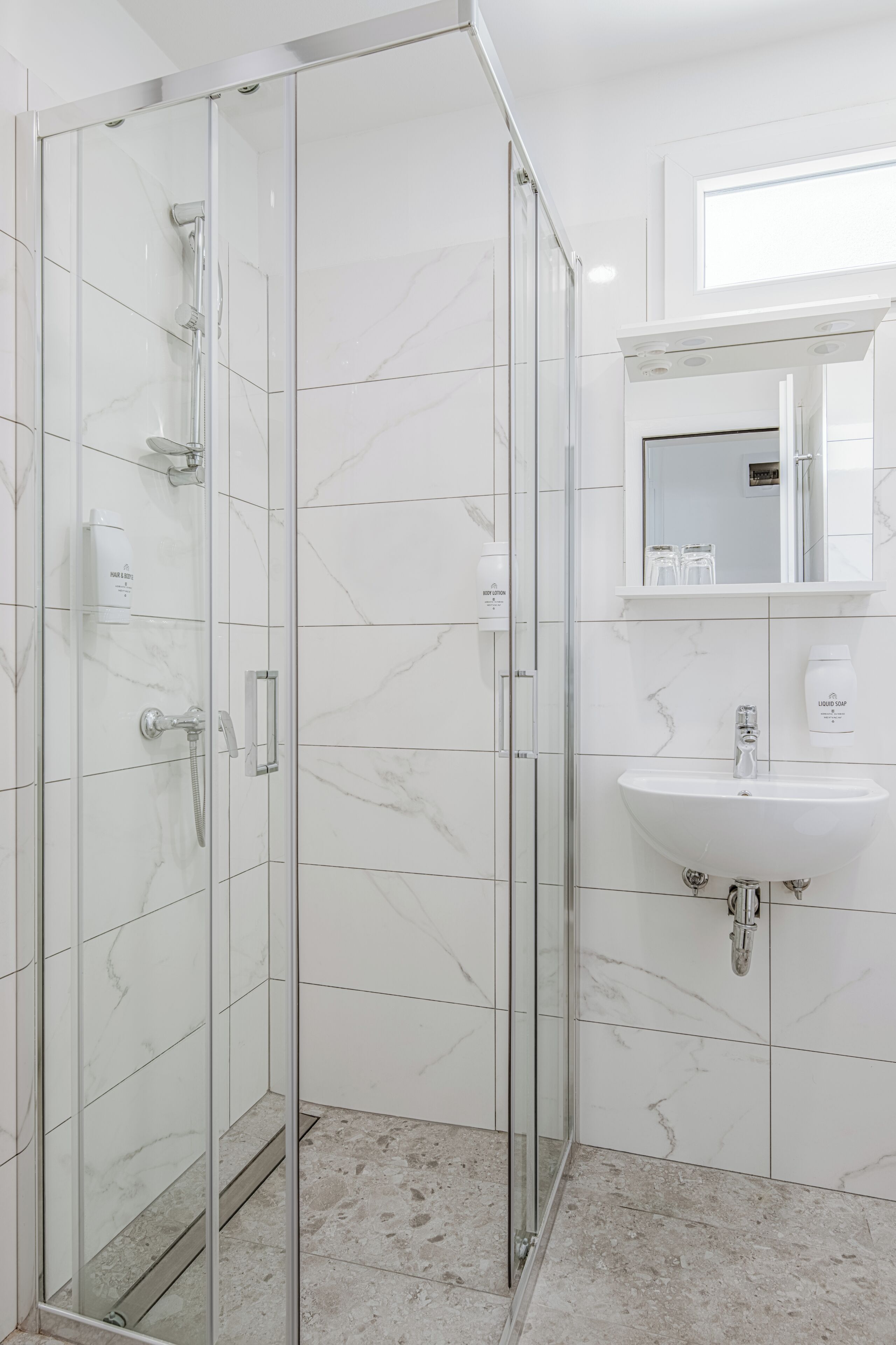 A modern bathroom with a glass-enclosed shower, white marble tiles, a sink, and a mirror above it.