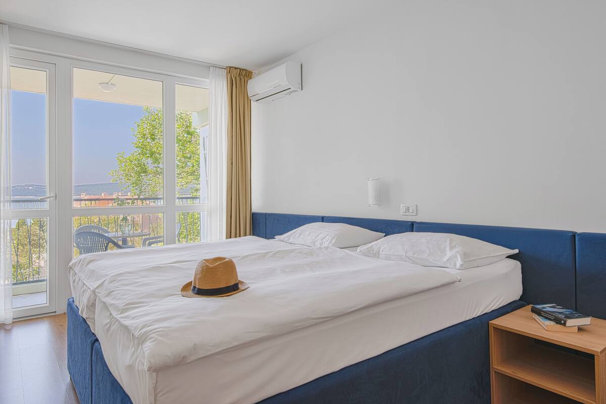 A hotel room with a large bed, a nightstand, an air conditioning unit, and a view of the outdoors.