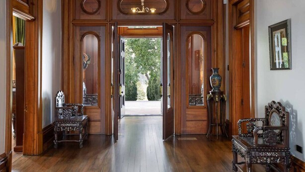 Una imponente entrada de madera con candelabro, dos sillones ornamentados y un jarrón al lado derecho.