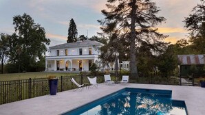 Una piscina con sillones de descanso, una casa grande con jardín y una cerca.