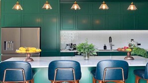 Cocina moderna con gabinetes verdes, refrigerador de acero inoxidable, una isla de mármol con taburetes altos e iluminación colgante.