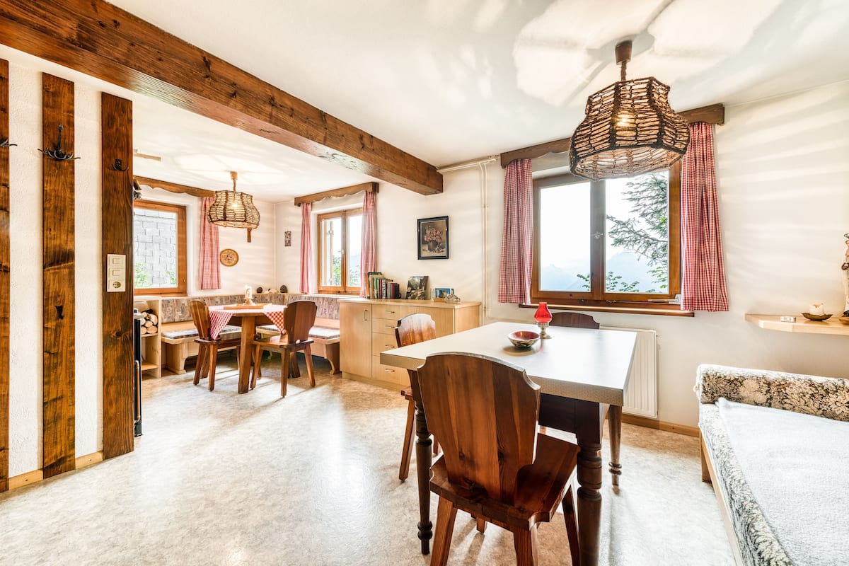 A cozy kitchen with wooden beams, a dining table, chairs, and a large window with curtains.