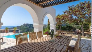 A wooden table and chairs under a pergola with a view of the sea and a hill. - Fiora Paradise Villa Resort (kythira)