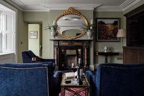 Una habitación con dos sillones azules, una chimenea con un espejo decorativo encima y un cuadro en la pared.
