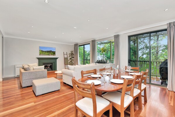 A spacious living area with a fireplace, a dining table set for six, and large windows offering a view of greenery.