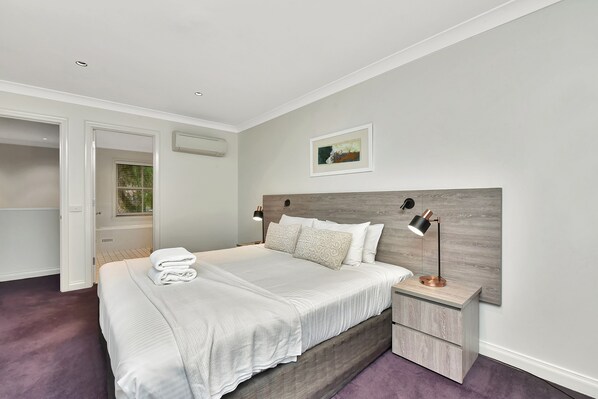 A bedroom with a bed, bedside tables, a nightstand, a wall-mounted air conditioner, a framed picture, and a door leading to another room.