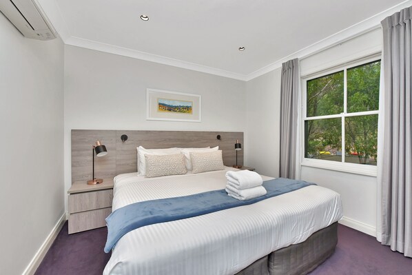 A hotel room with a large bed, a nightstand, a lamp, a painting on the wall, and a window with curtains.