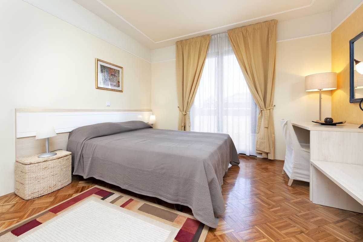 A bedroom with a bed, a bedside lamp, a bedside table with a lamp, a window with curtains, and a wall-mounted picture.