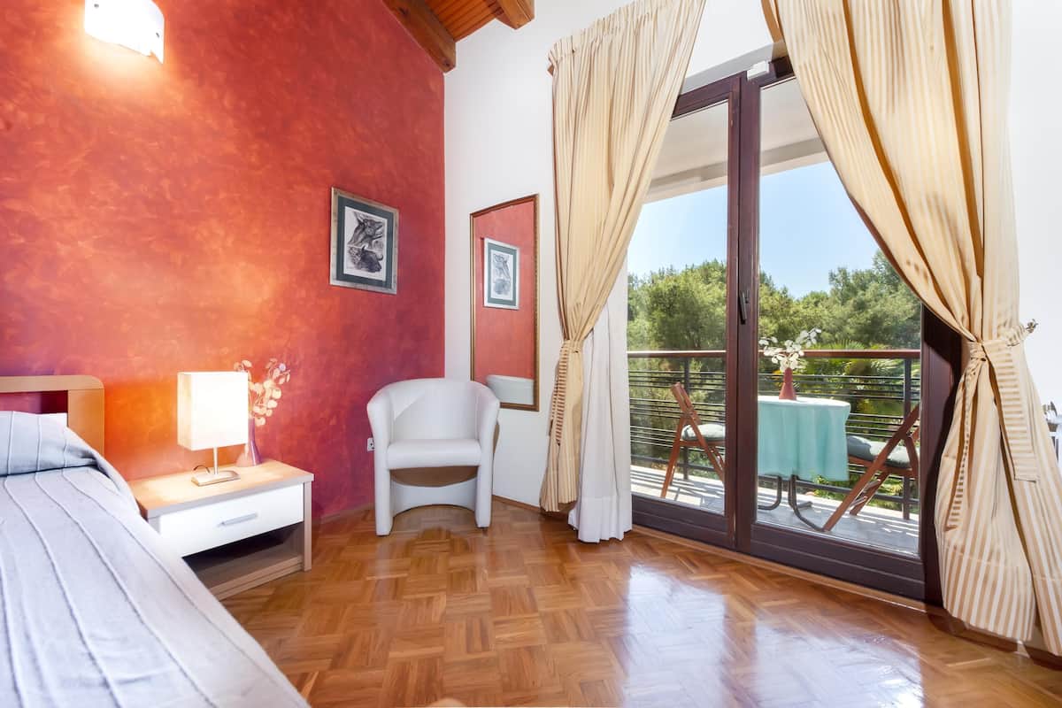 A bedroom with a wooden floor, a bed, a nightstand with a lamp, a chair, and a balcony with a table and chairs.