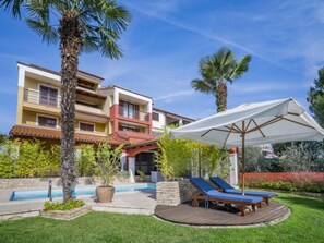Ein mehrstöckiges Gebäude mit Swimmingpool, Liegestühlen und einem überdachten Bereich mit Sonnenschirm.