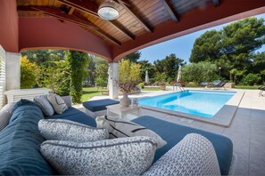 A covered outdoor seating area with a pool, lounge chairs, and a view of the surrounding greenery.