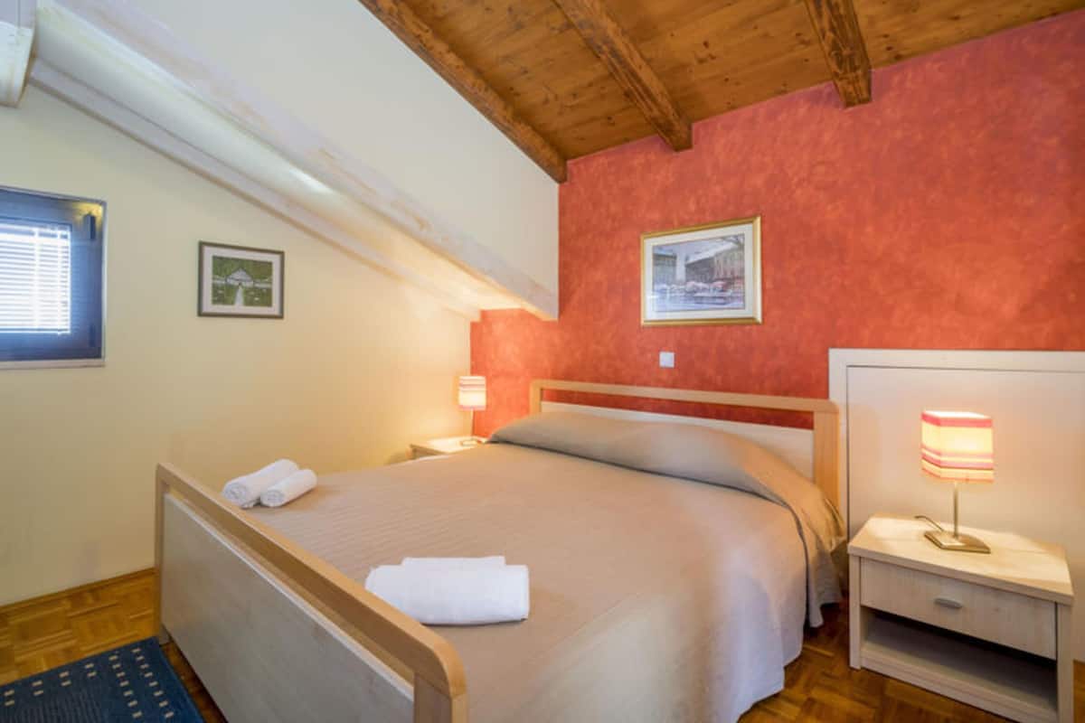A cozy bedroom with a wooden ceiling, a bed with a white headboard, two bedside tables with lamps, and two framed pictures on the walls.